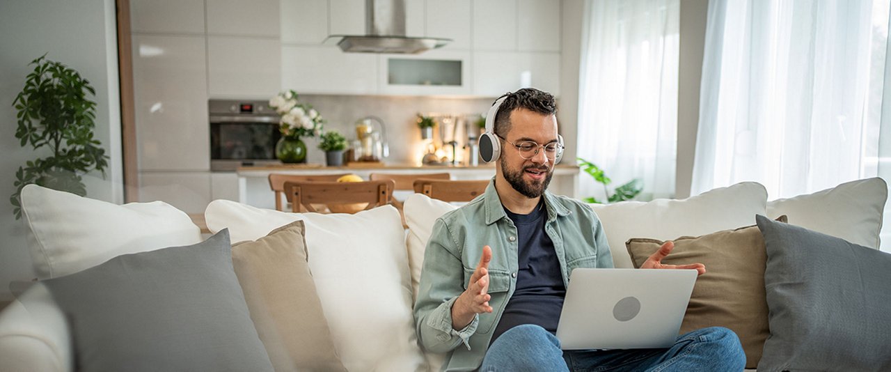A man wearing headphones sits on a couch, focused on his laptop in a cozy living room setting.