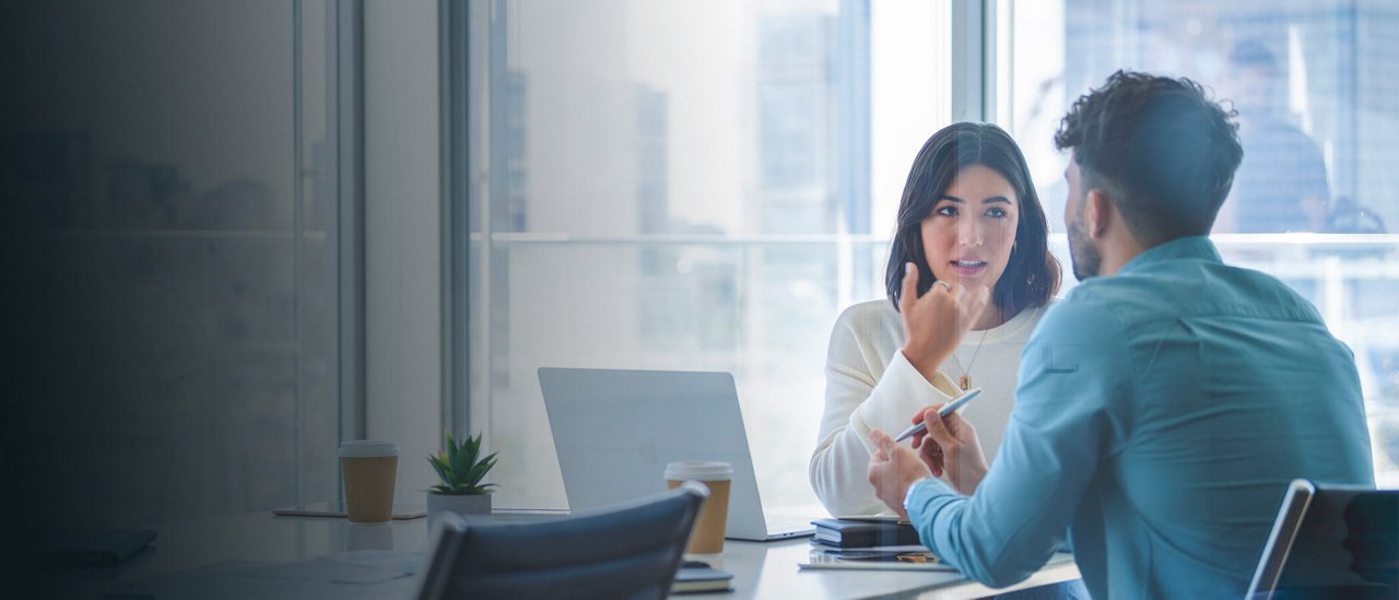 A man and woman are seated at a table, focused on a laptop, engaged in a collaborative discussion.