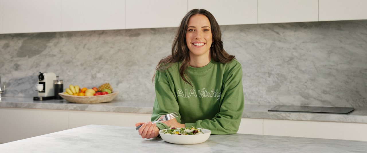 A woman in a green shirt sits at a table, enjoying a bowl of salad.
