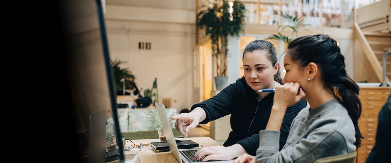 Two women collaborating on a laptop in a modern office setting, focused on their work.