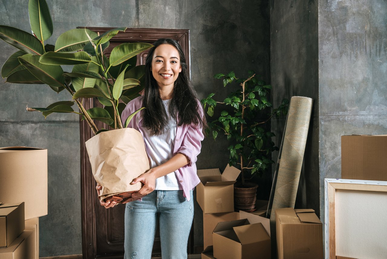 一位年輕女性剛剛搬入新房，抱著一盆植物，周圍堆著紙箱。 