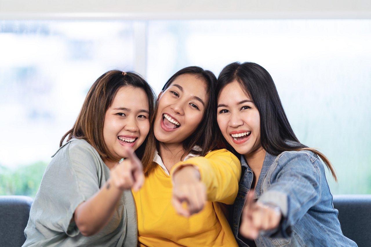 Three smiling Asian women point at the camera.