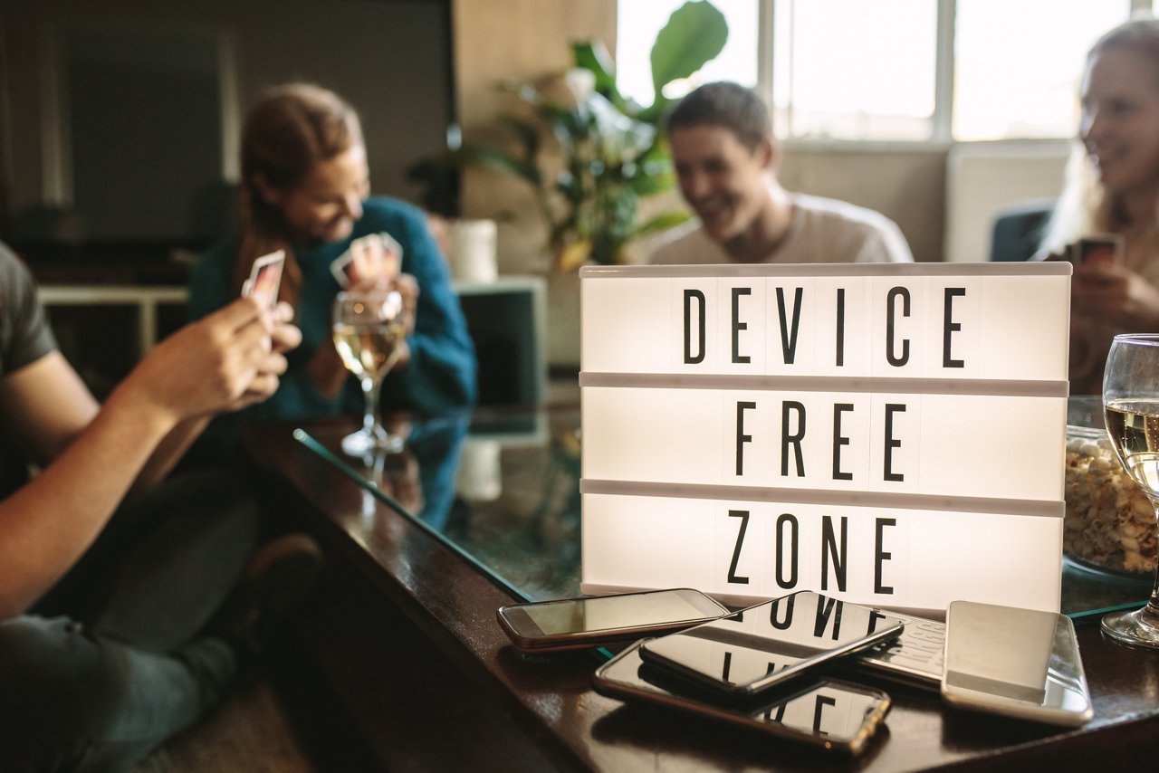 Pile of mobile phones in front of a "device free zone" sign on a table, with a group of people conversing in the background.