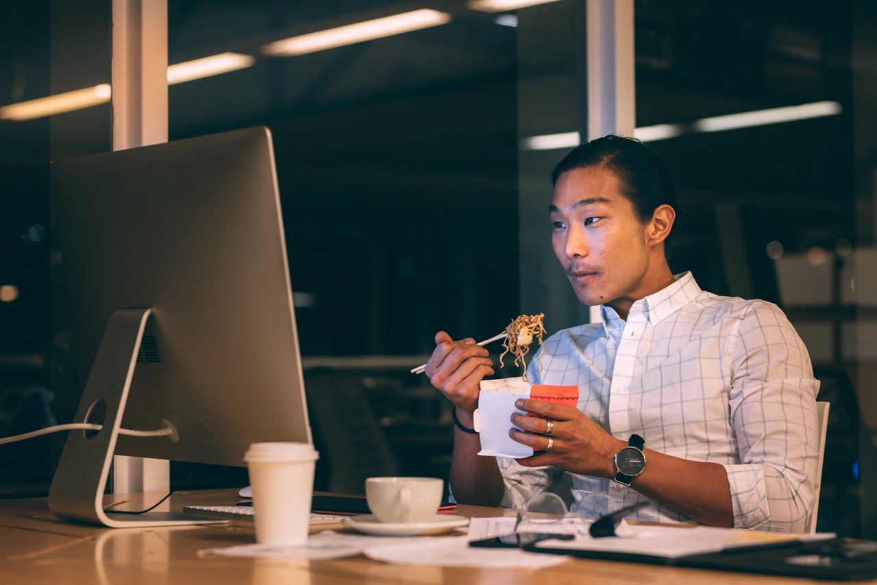 一位亞洲男子在辦公桌前對著電腦吃即食麵。