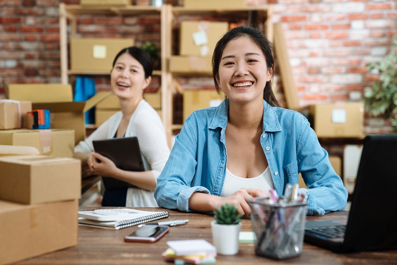 Two young women surrounded by boxes smile at the camera.