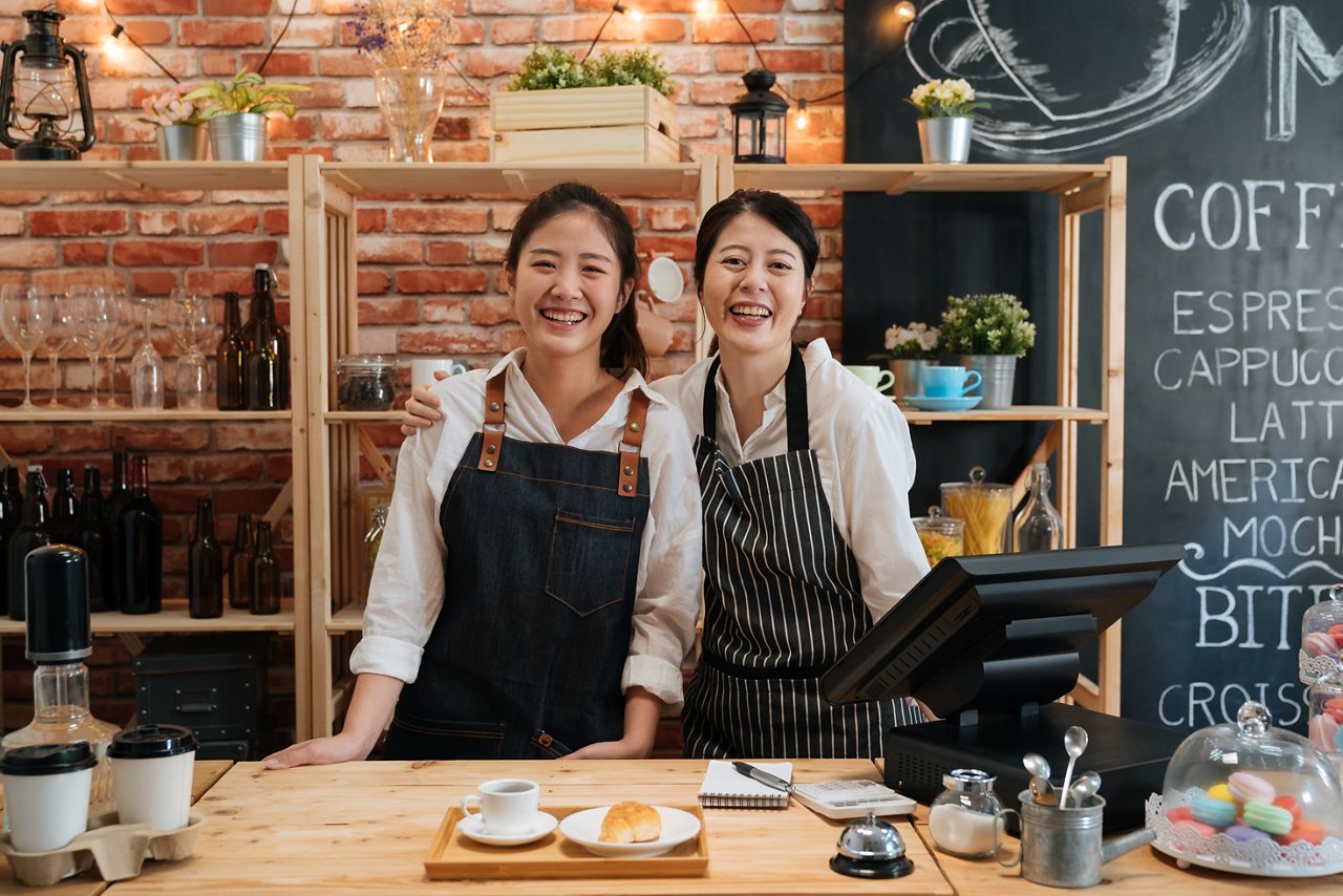 Two Asian women in aprons stand behind the cashier at their café. 