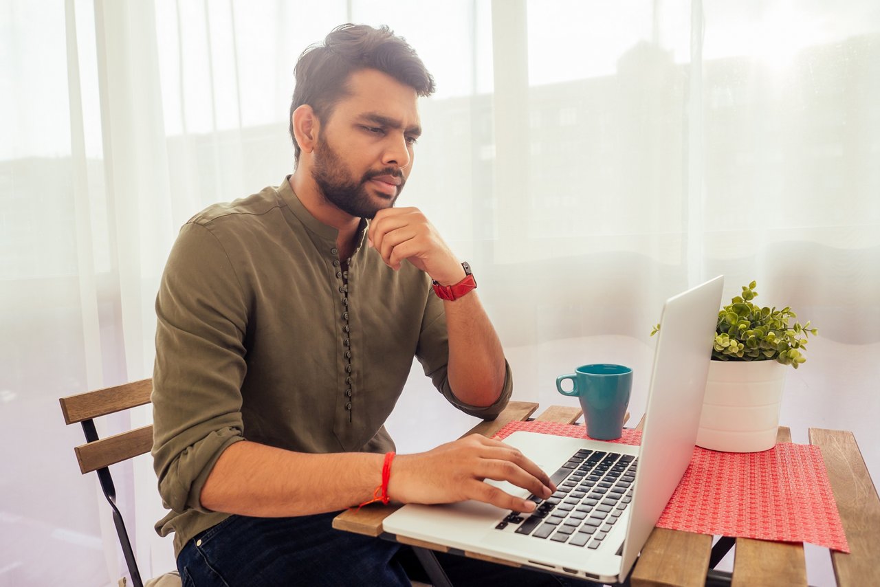 Man working on a laptop and drinking coffee from blue cup at his home.