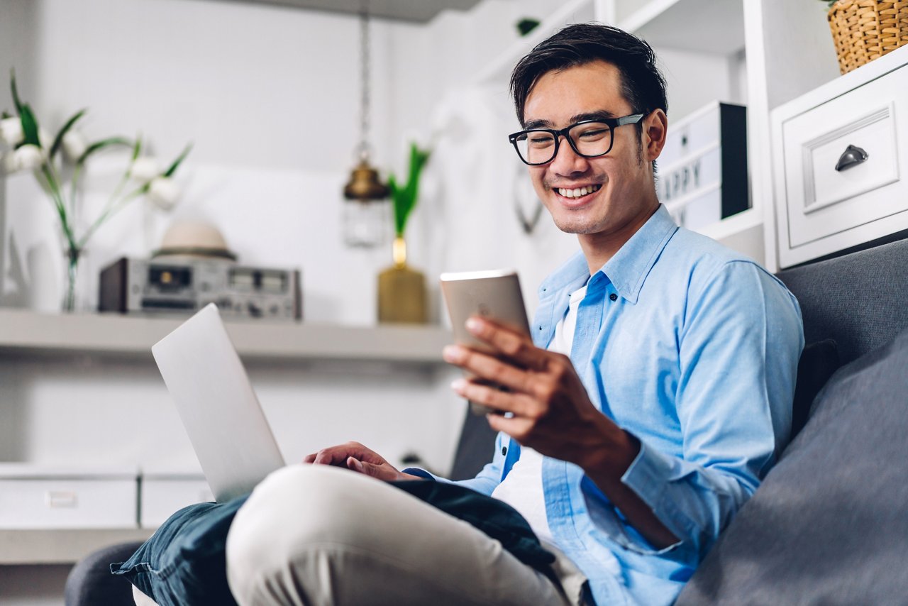 Young Asian male with a laptop on his lap checks his mobile phone