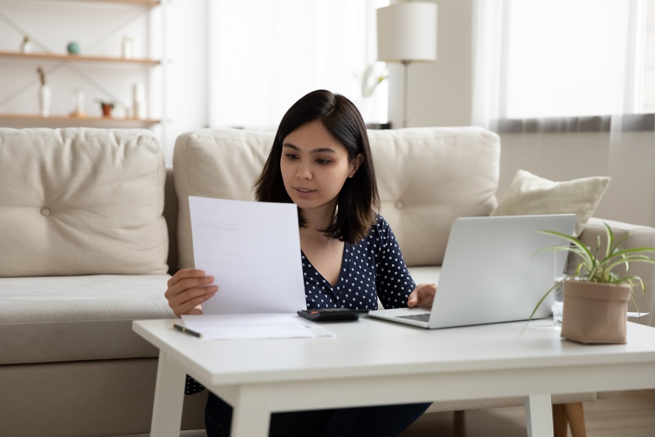一位年輕女性邊用筆記本電腦打字邊看著一張紙。  