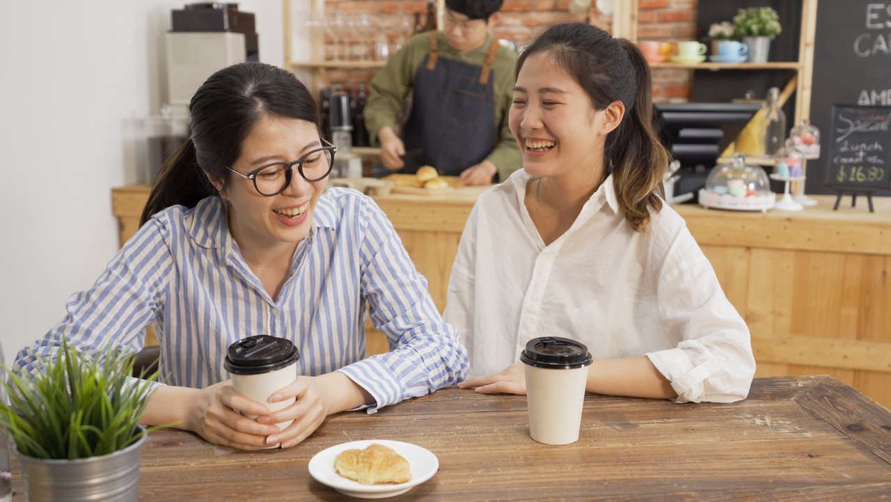 Two female co-workers chat while waiting for meals at a coffee shop.