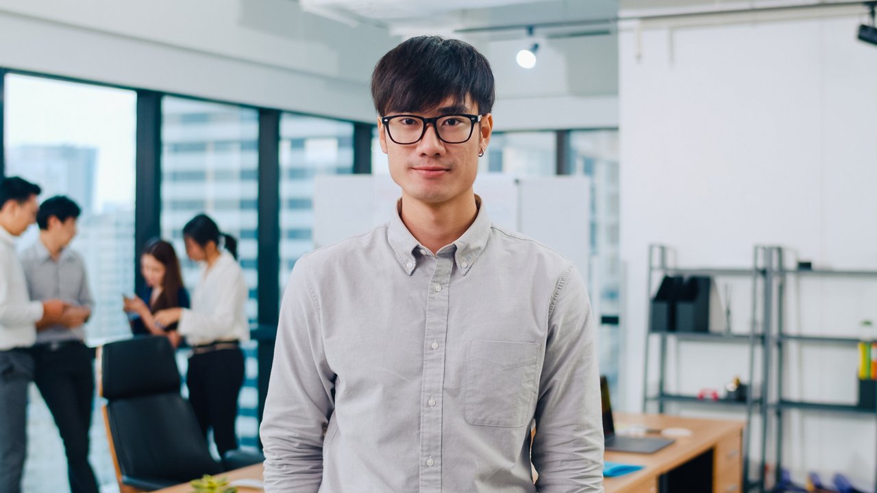 Asian businessman looks at the camera while in a modern office with colleagues in the background.
