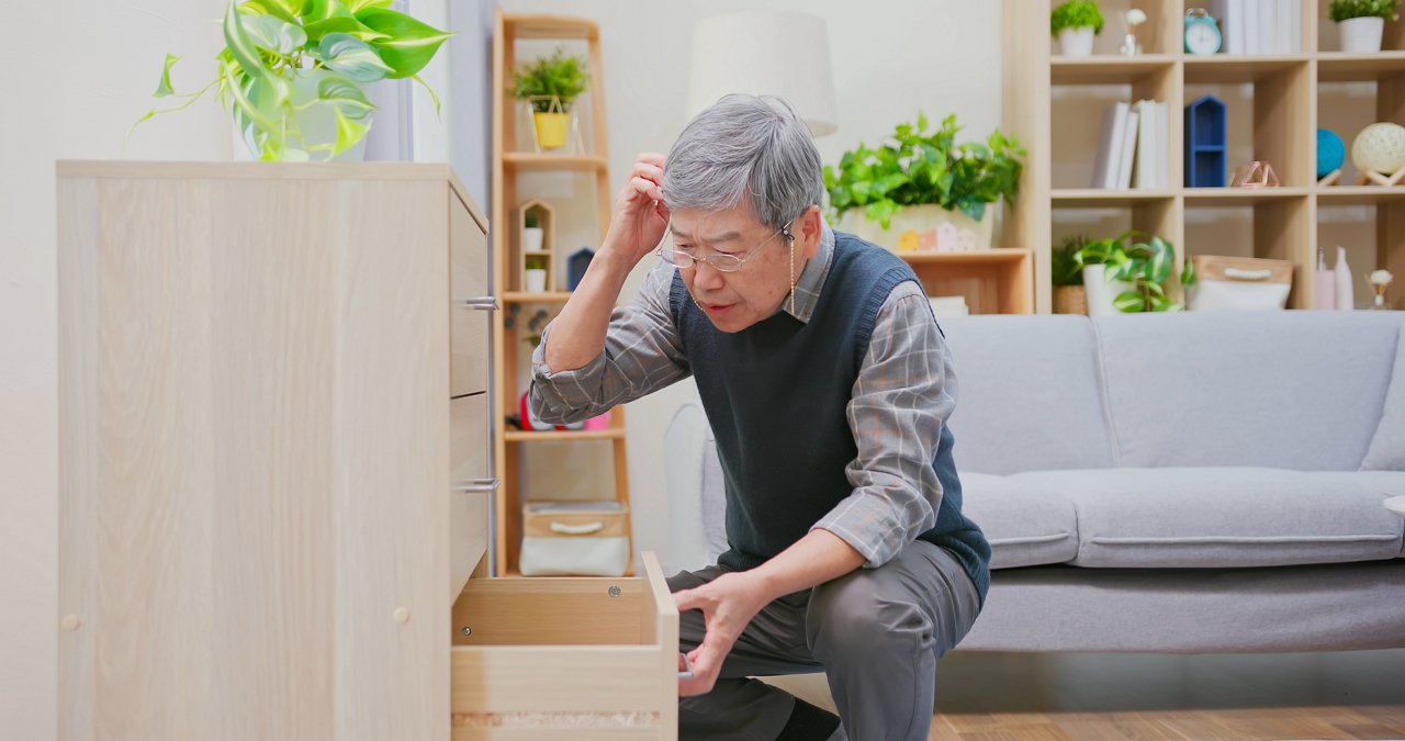 Senior Asian man scratches his head as he looks for something in a drawe