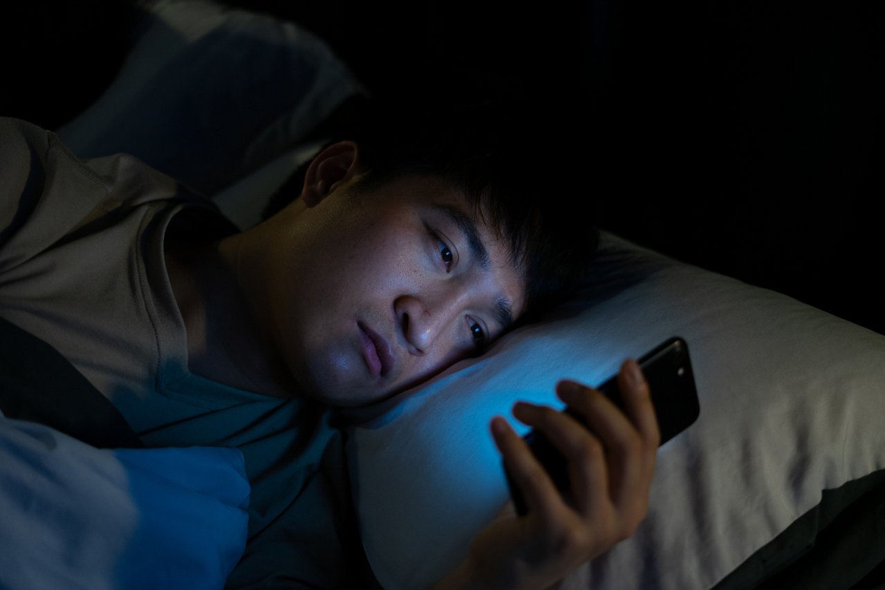 一位年輕的亞裔男子深夜盯著智能手機，手機藍光影響了他的睡眠。 