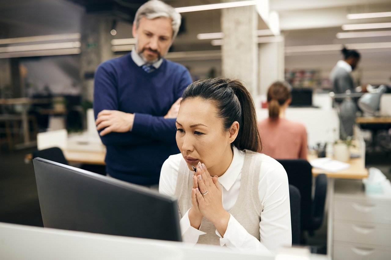 Asian woman looks at her laptop while her boss stands behind her.