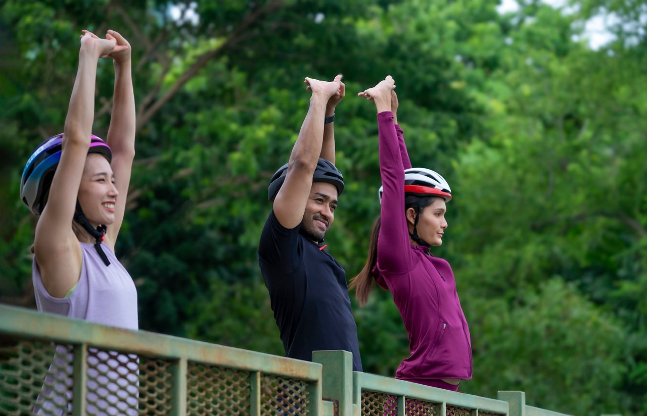 Three friends wearing helmets in a park stretch their arms over their heads.
