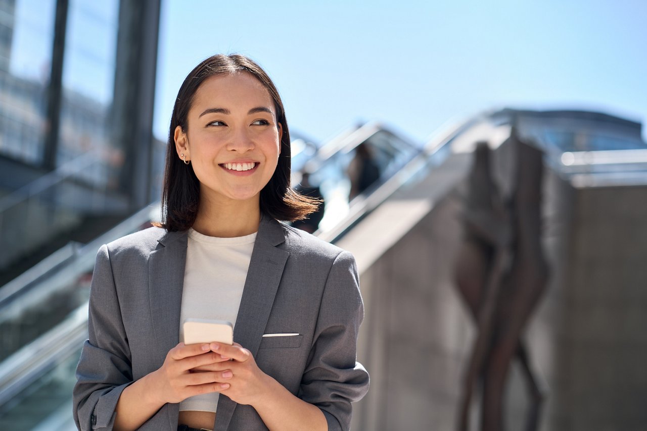 一位年輕的亞裔職場女性微笑著手持智能手機站在室外。 