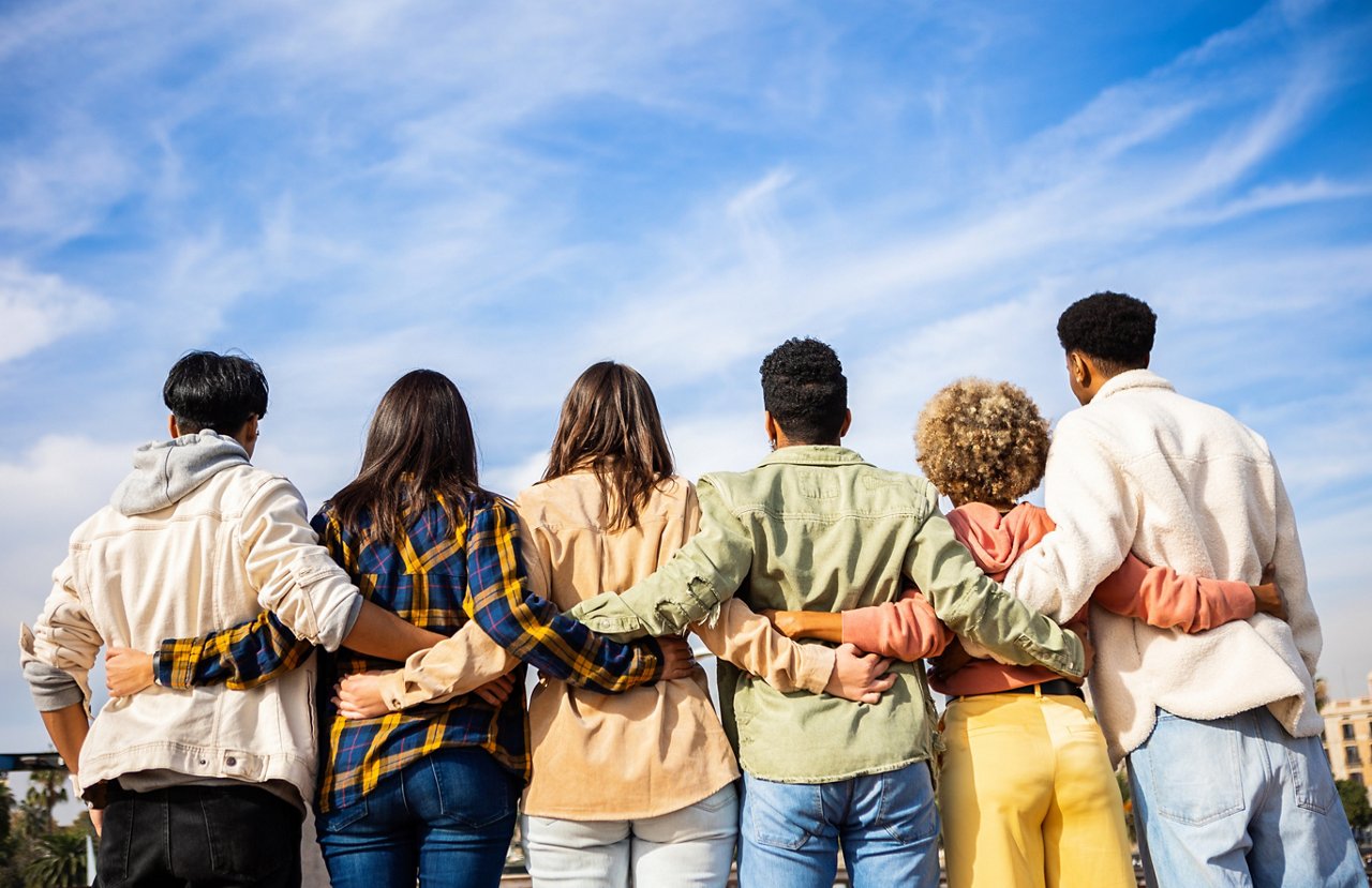 Rear view of a group of friends standing side by side while hugging each other outdoors.