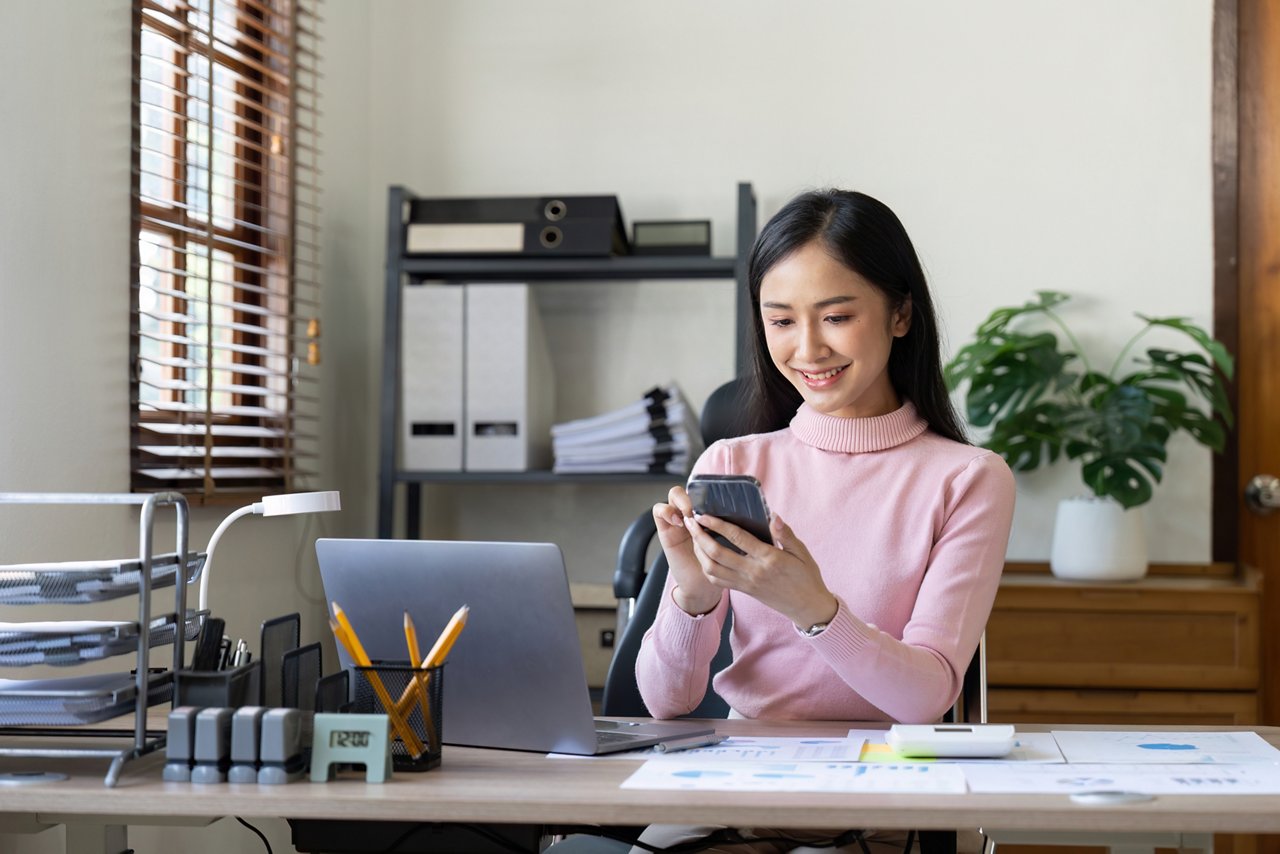 Young Asian woman checks her phone at her work desk.