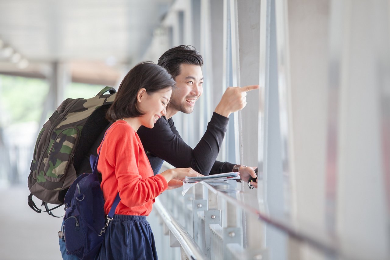 Backpack-carrying Asian couple checks the travel guide and looks out at an overpass.