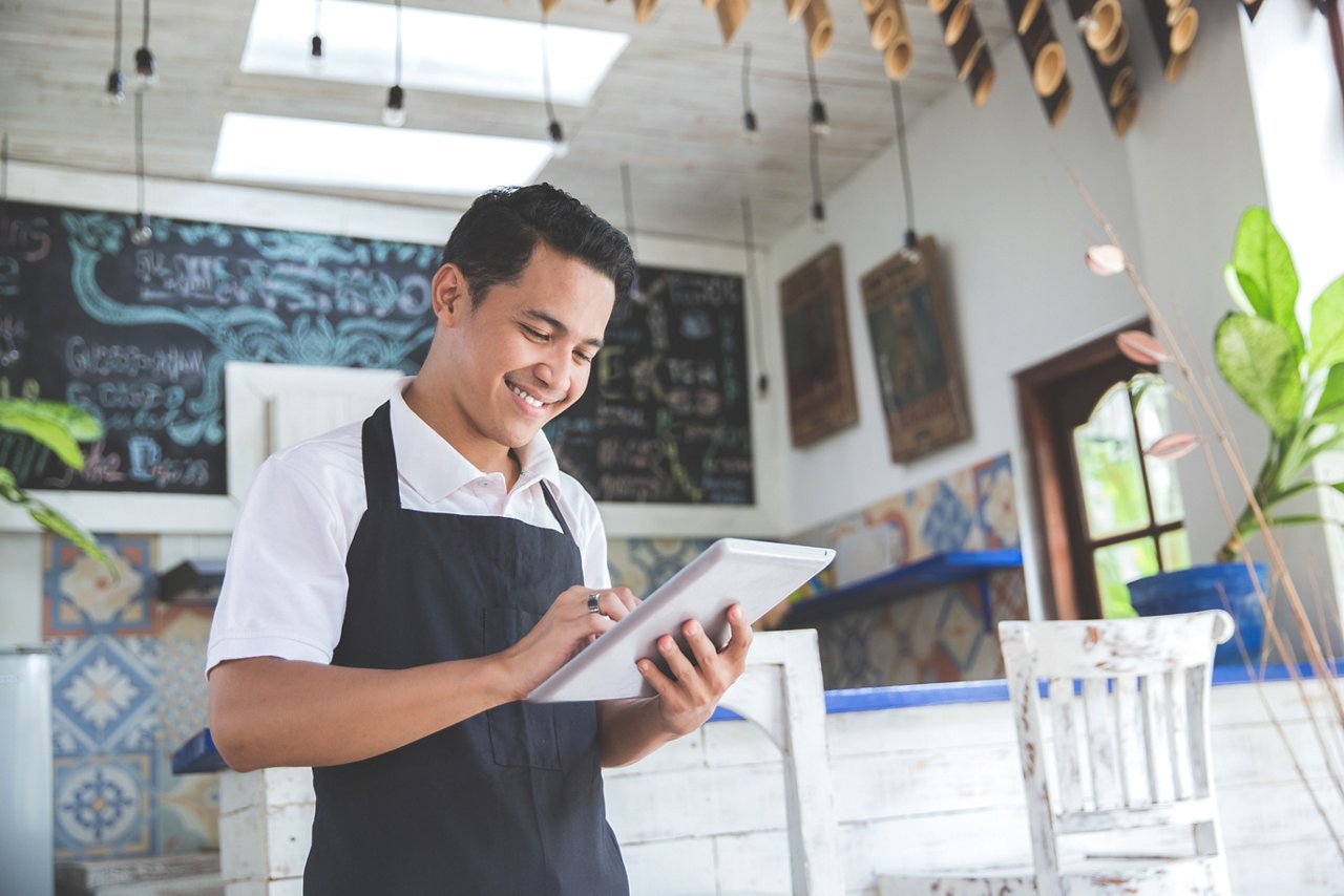 A young male cafe owner holds a tablet to check the finances of his business.