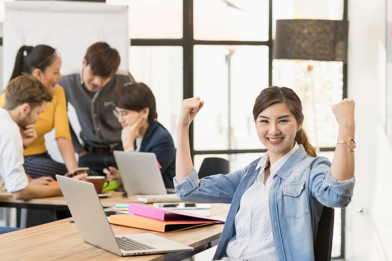 Smiling Asian woman with her arms up expressing victory at her workplace.