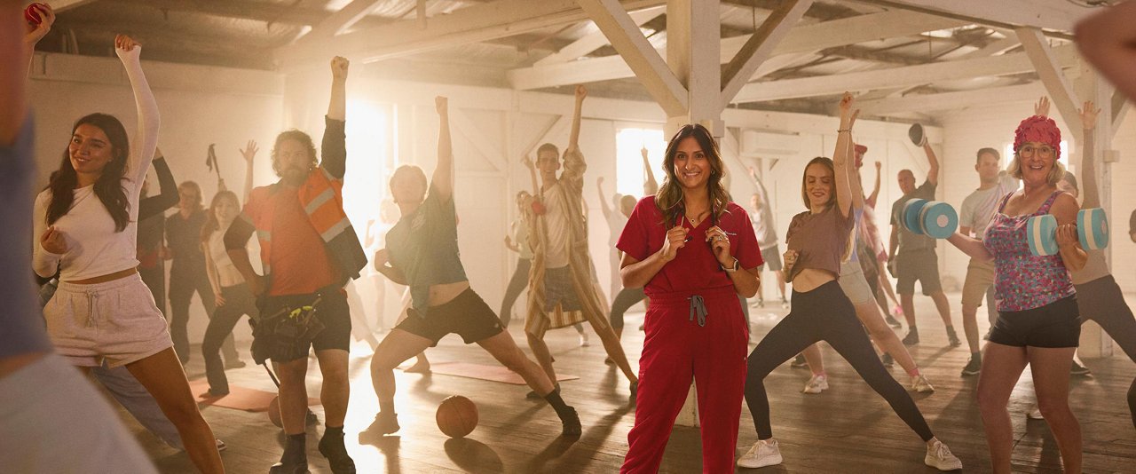 Dr Preeya Alexander standing in front of a diverse group of individuals engaged in a lively dance session inside a spacious gymnasium.