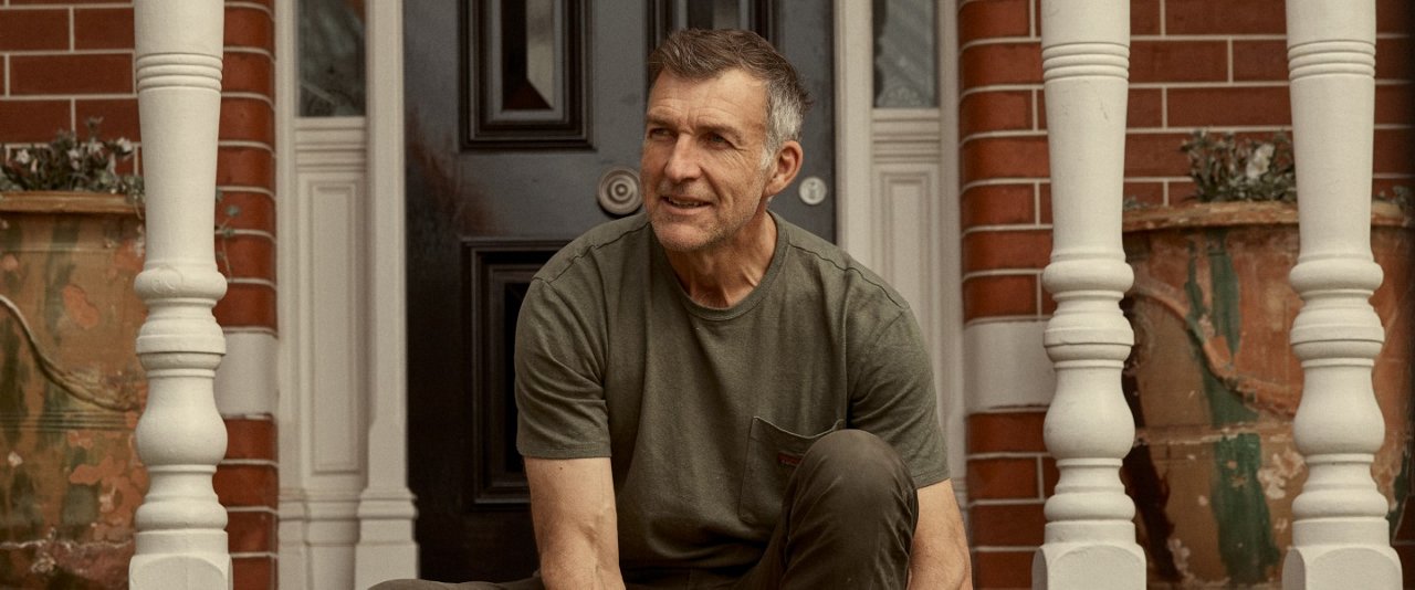 A man seated on the ground beside a house, appearing relaxed in a tranquil outdoor setting.