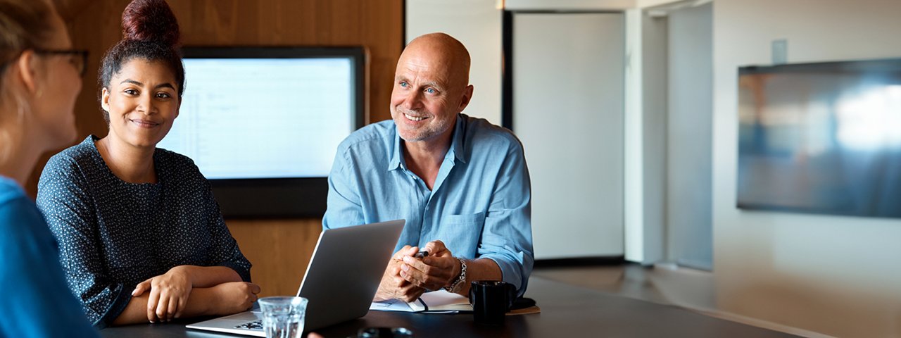 Business meeting in progress with a senior man and a young woman smiling at a colleague, sitting at a table with a laptop open.