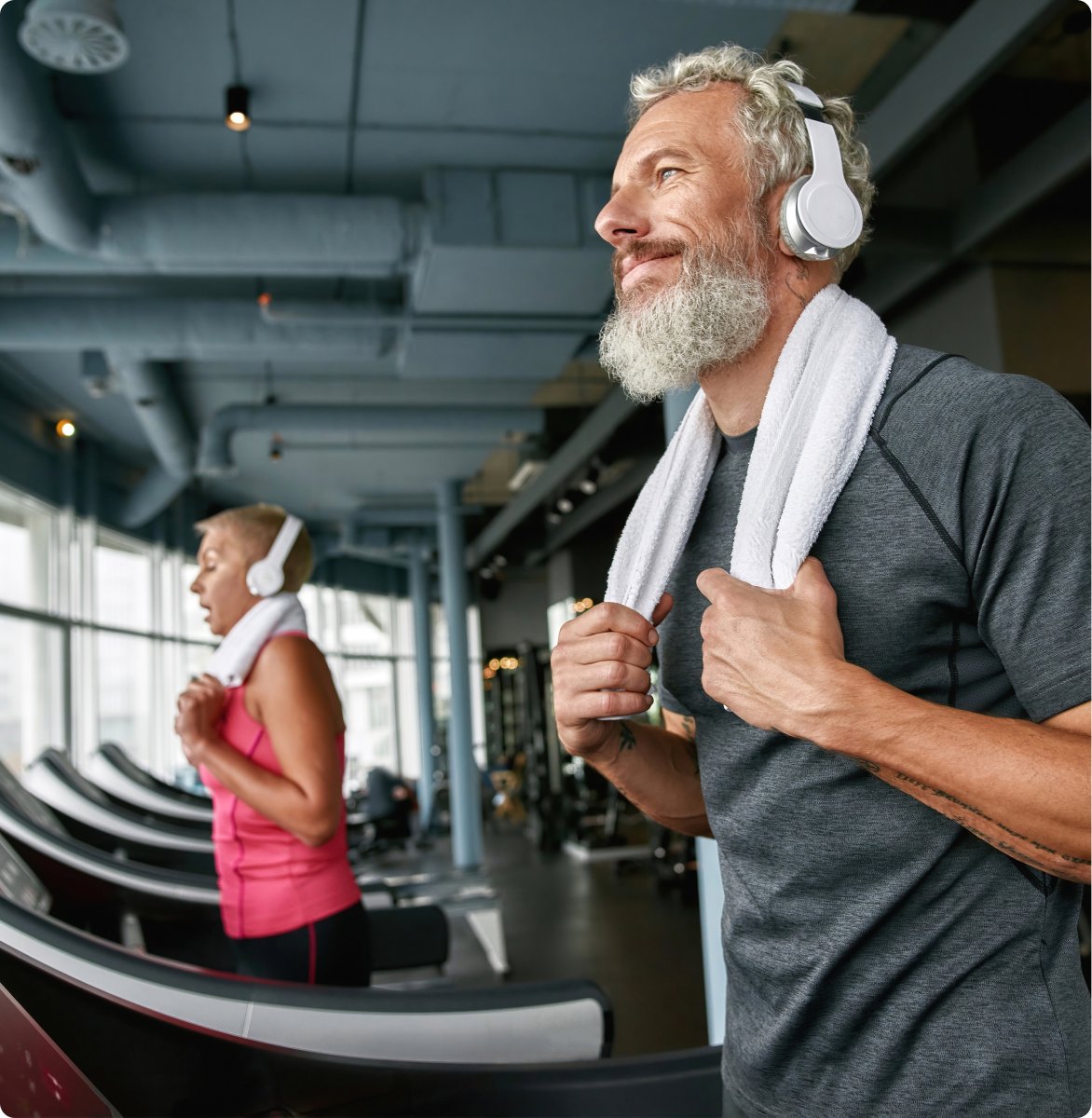 A bearded man wearing headphones runs on a treadmill, focused on his workout in a gym setting.