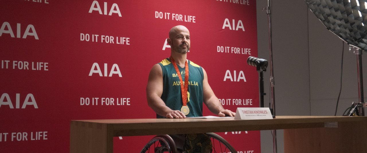 A man in a wheelchair sits at a table, speaking into a microphone during a discussion or presentation.