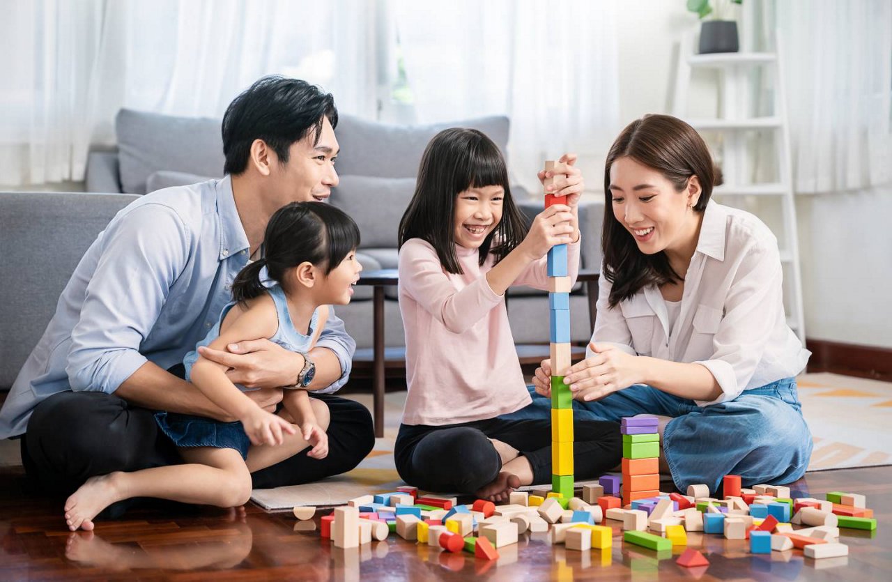 一對亞洲夫婦正帶著兩個年幼的女兒在客廳地板上玩彩色積木。   