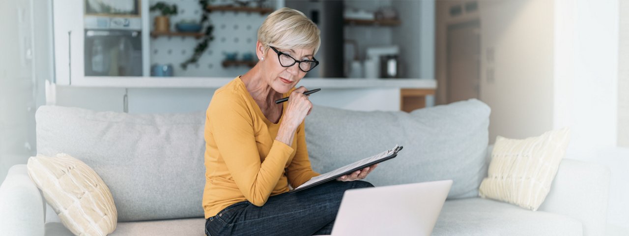 A woman sitting on a couch, using a laptop and holding a pen, focused on her work.