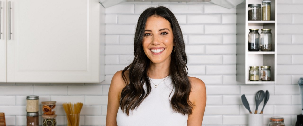 A woman wearing a white top stands in a modern kitchen, surrounded by cooking utensils and appliances.