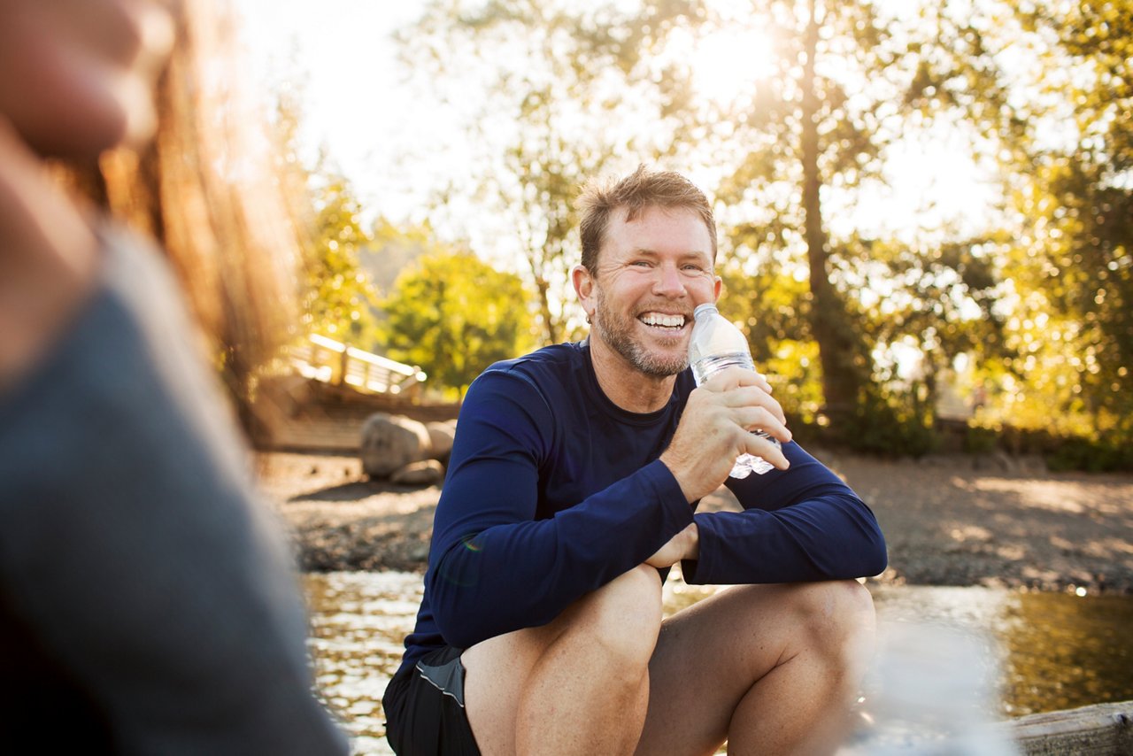 A man seated on a dock, holding a water bottle, with serene water in the background.