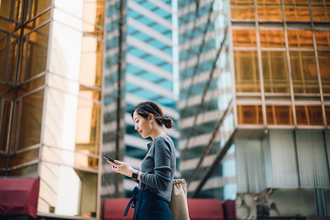 Low angle portrait of young Asian businesswoman checking emails on smartphone outside office buildings in financial district. With contemporary corporate skyscrapers in background. Business on the go. Making business connections throughout the city