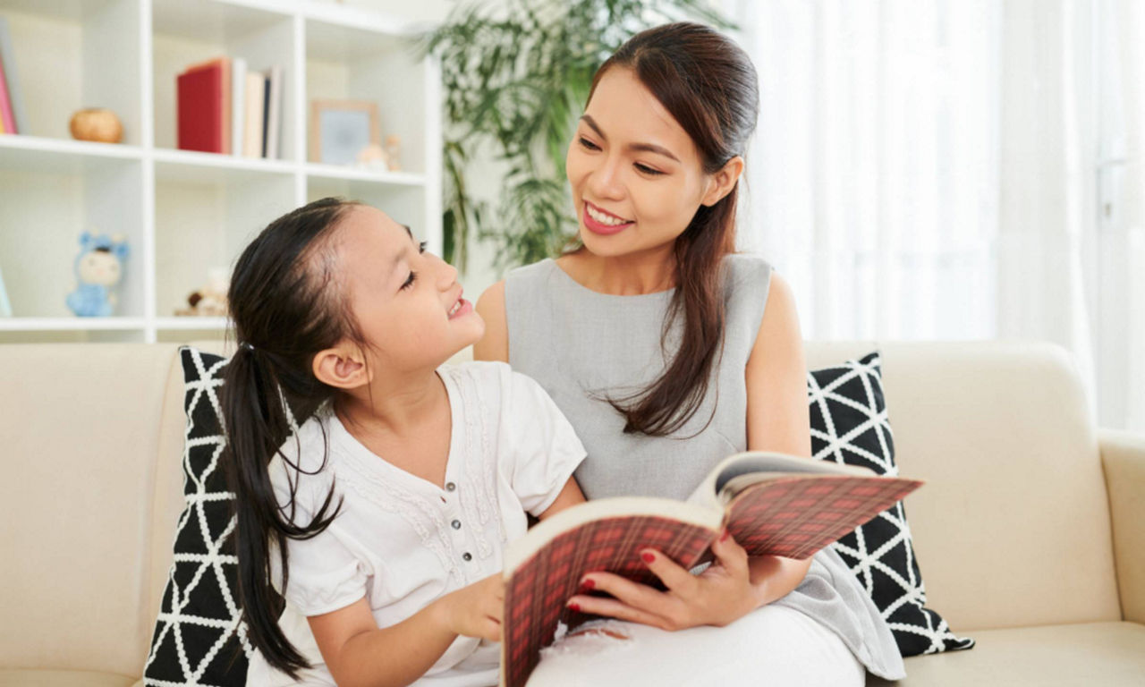 Young Asian mother smiling and reading a book together with her little daughter while they sit on sofa at home 