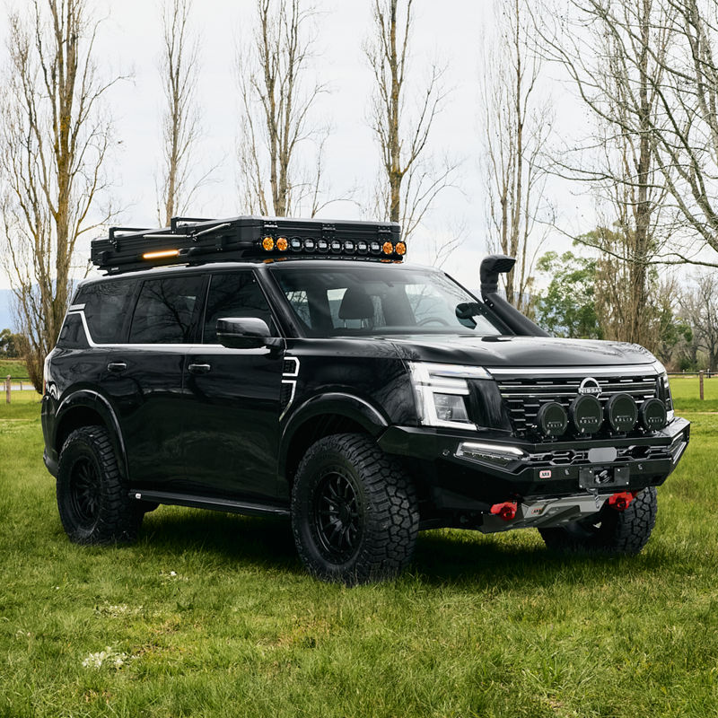 Front three-quarter view of a Nissan Patrol Y63 fitted with an ARB bull bar, driving lights, roof rack, snorkel and under-vehicle protection, parked on grass.