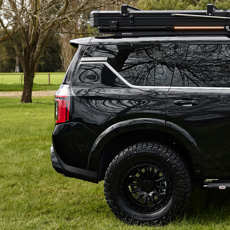 Close-up rear side view of a Nissan Patrol Y63 showing ARB roof rack, awning, all-terrain tyre and rear quarter panel, parked on grass.