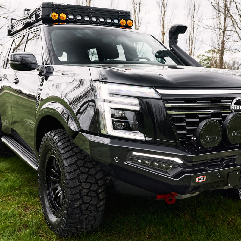 Close-up front three-quarter view of a Nissan Patrol Y63 showing an ARB bull bar, driving lights, roof rack, snorkel and all-terrain tyres, parked on grass.