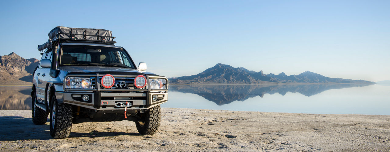 Front three quarter view of a Toyota LandCruiser 100 Series fitted with an ARB bull bar, driving lights, roof rack system, snorkel and winch, parked on a salt flat with mountains reflected in the water.
