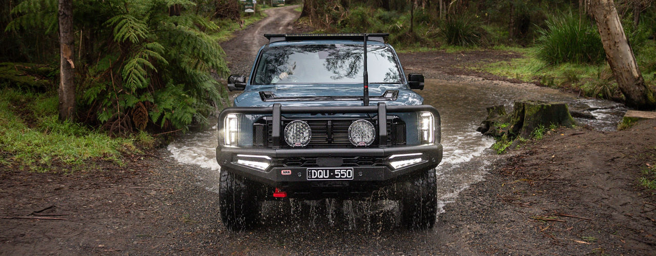 Front view of a Kia Tasman fitted with an ARB bull bar, Intensity driving lights and roof rack, crossing a shallow creek in dense forest.