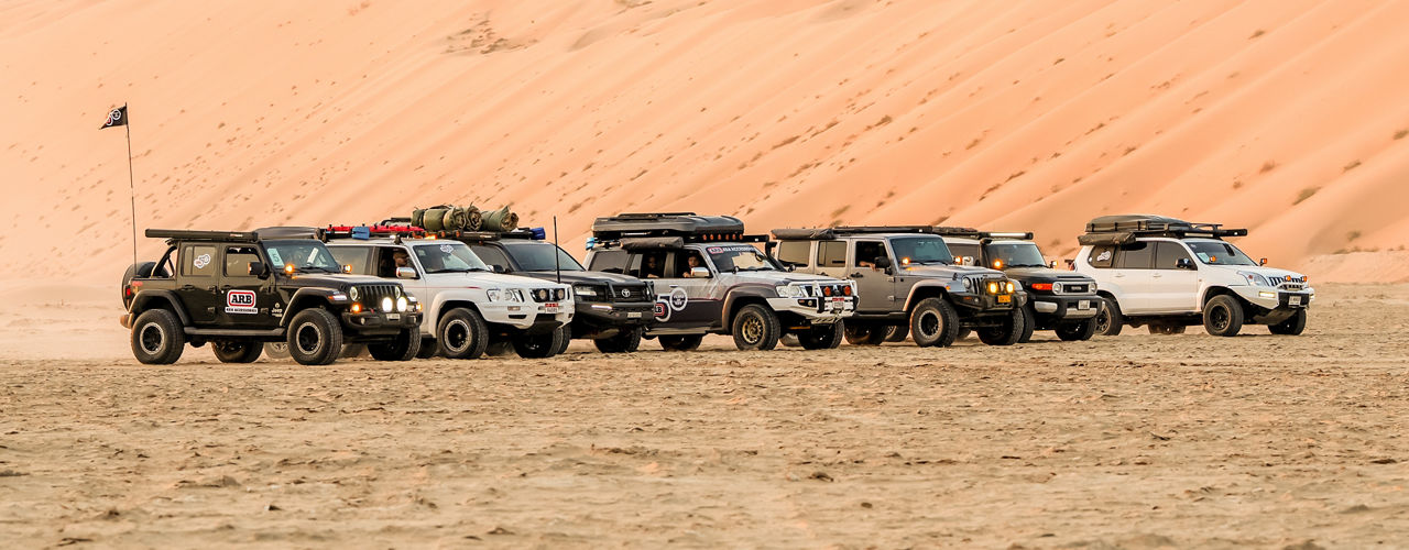 Line-up of ARB equipped 4WD vehicles parked in front of sand dunes in a desert environment.