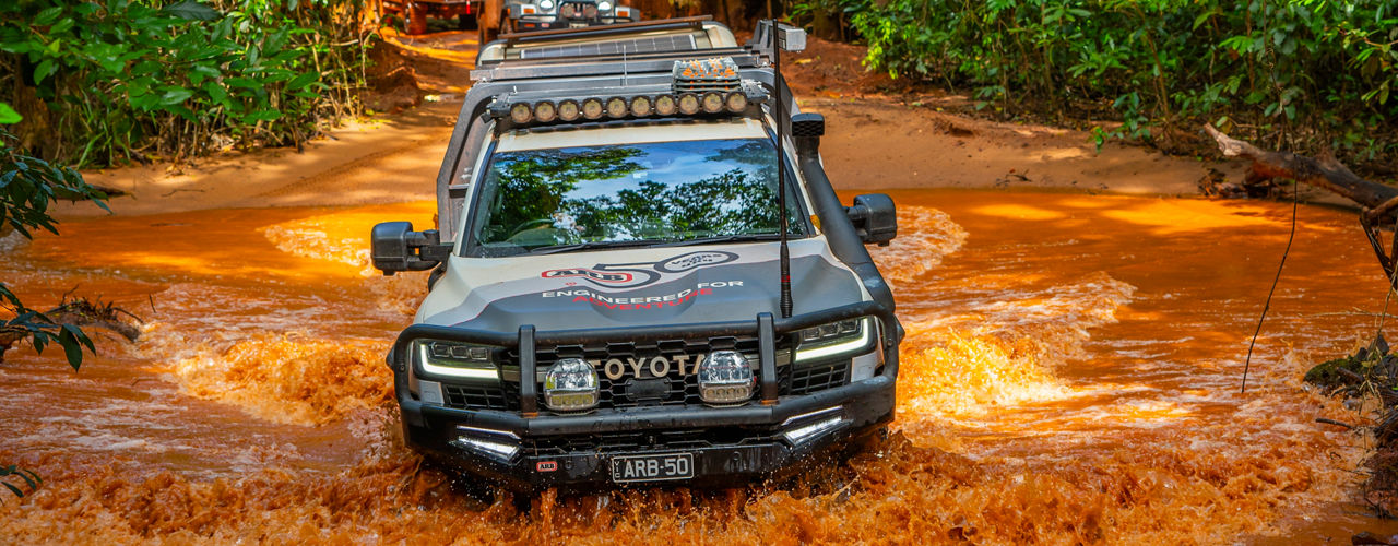 ARB equipped Toyota vehicle crossing a muddy creek in a dense forest, with other vehicles visible in the background.