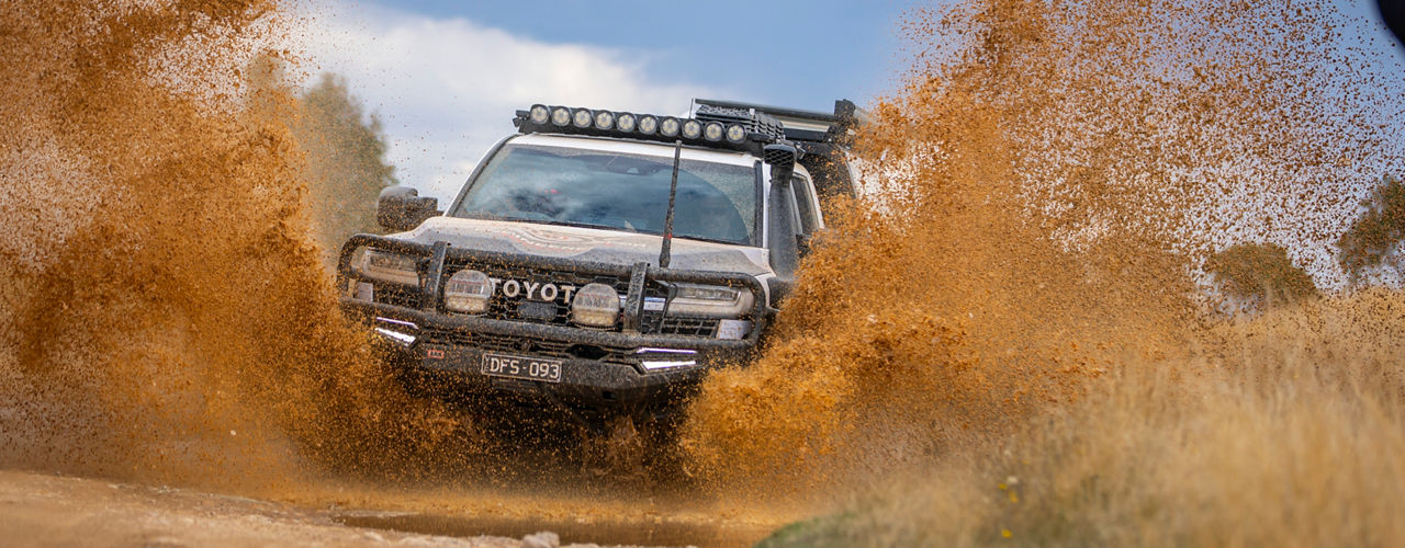 Front view of an ARB equipped 4WD vehicle driving through a muddy water crossing, with mud spraying around the vehicle and a roof rack system visible above the cab.