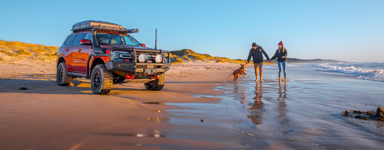 Couple with a pet dog walking at a beach beside a Ford Everest.