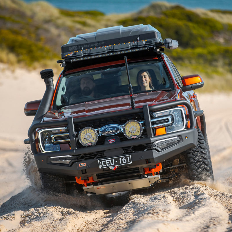 Ford Everest fitted with an ARB bull bar, driving lights, roof rack system and rooftop cargo box driving through deep sand, with two occupants visible inside the vehicle.