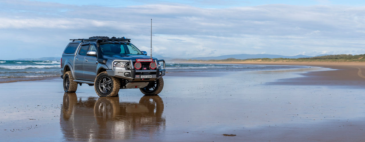 Wide view of a Ford Ranger PXIII fitted with an ARB bull bar, driving lights, snorkel, canopy, roof rack and under-vehicle protection, parked on a beach shoreline.