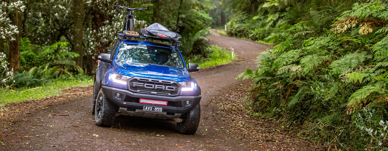 Ford Ranger Raptor driving along a forest track, fitted with a roof rack system carrying bicycles and outdoor gear.