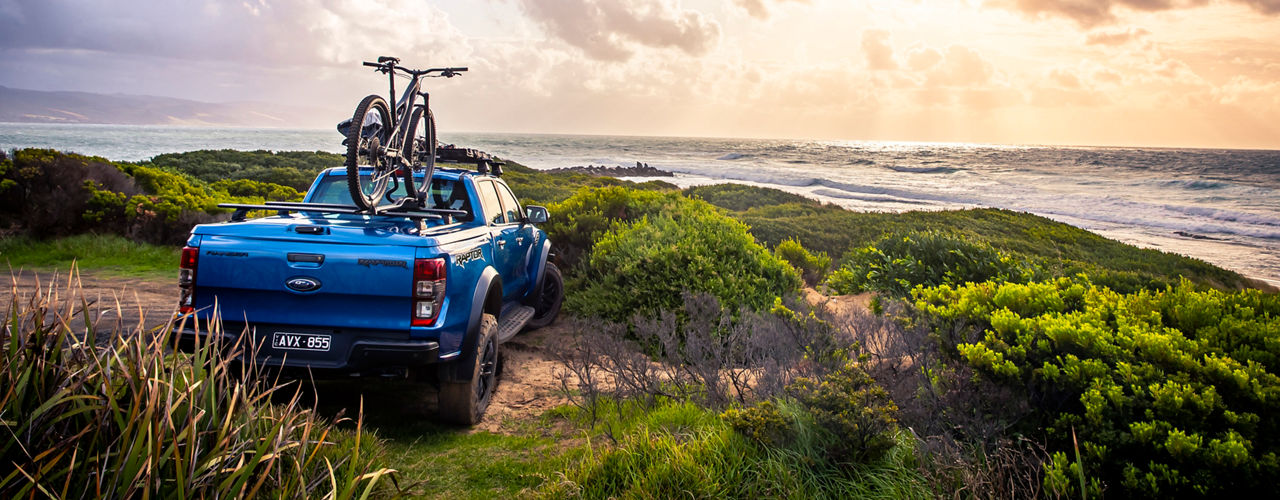 Ford Ranger Raptor parked on a coastal track overlooking the ocean at sunset, fitted with a roof rack system carrying a bicycle.