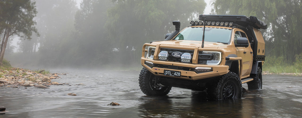 Ford Ranger Super Duty fitted with a canopy, roof rack system, awning and driving lights during a creek crossing in a misty forest setting.